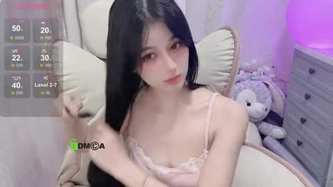 Media: A video of an East Asian woman with long black hair and pale skin, wearing a pink lace bralette, sitting on a cream-colored cushioned chair. Background includes a stuffed toy and a purple-lit room.