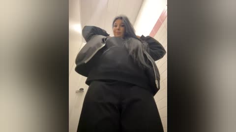 Media: Video of a plus-sized woman with long black hair, wearing a black jacket and pants, standing in a public restroom with tiled walls and fluorescent lights.