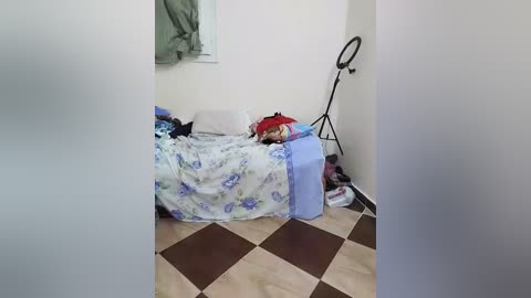 Media: Video of a cluttered bedroom with a messy bed covered in a blue floral duvet, clothes, and a camera tripod in the corner.