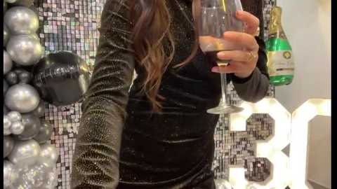 Media: Video of a woman in a black glitter dress holding a glass of champagne. Background features shiny silver and black decor, including a wall of sequined tiles and round white lights.