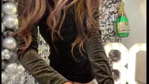 Media: A video of a woman with long, wavy brown hair, wearing a black glittery top, standing in front of a glitter wall and a glowing white hexagonal lamp.