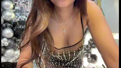 Media: Video of a woman with long, wavy brown hair, wearing a sheer black lace dress with a plunging neckline, and a delicate gold necklace. Background features a mosaic wall and silver ornaments.