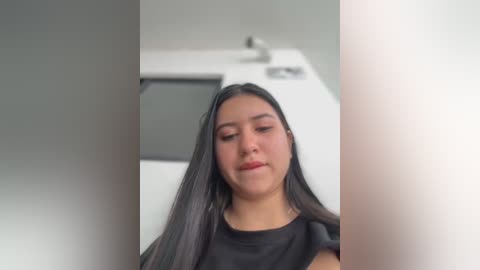 Media: Video of a young Latina woman with long, straight, black hair, wearing a black sleeveless top, standing in a modern, minimalist kitchen with white counters and stainless steel appliances.