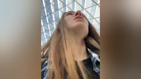 Media: A video of a young woman with long, light brown hair, wearing a plaid shirt, standing in a modern building with a grid-like glass ceiling. Her head is tilted back, eyes closed, and her expression is serene.