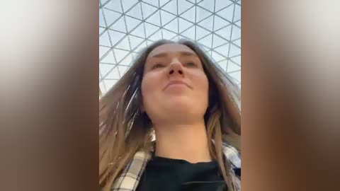 Media: Video of a young woman with long brown hair, wearing a black shirt and plaid shirt, standing under a geometric glass ceiling.