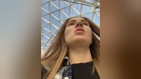 Media: Video of a young woman with long, straight brown hair, wearing a blue and white plaid shirt, viewed from below, with a modern glass and steel ceiling in the background.