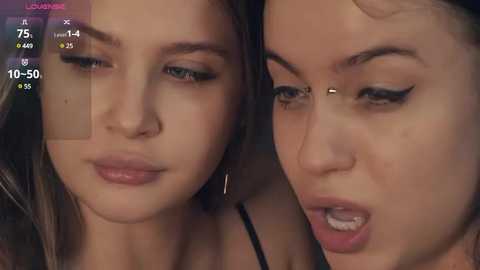 Media: Video of two young women with fair skin and long brown hair, one with a nose piercing, both wearing makeup, looking towards each other with serious expressions.