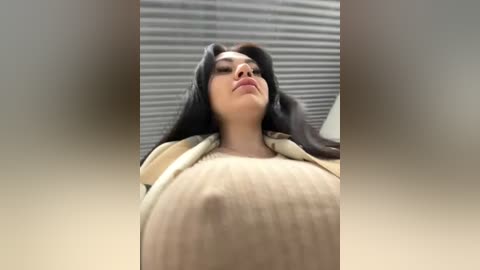 Media: A video of a woman with long black hair, wearing a beige sweater, standing in a room with horizontal blinds. She has a large, round belly, with visible nipples through the fabric.