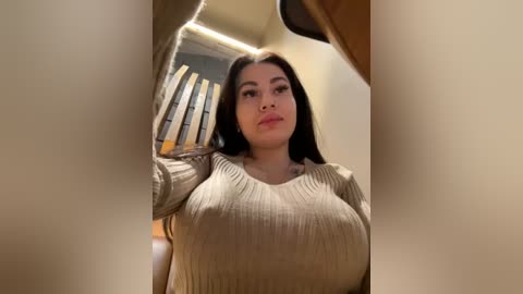 Media: Video of a young, curvy Latina woman with long black hair, wearing a beige ribbed sweater, posing in a modern, beige-toned room with wooden blinds and a soft light source.