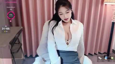 Media: A video of an East Asian woman with long black hair, wearing a white blouse with a deep neckline, seated in a room with pink curtains.