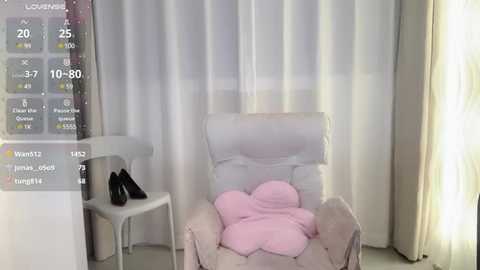 Media: Video of a modern, minimalistic bedroom with white curtains, a plush, light-colored armchair, and a small white table. A pink blanket is neatly folded on the chair, and a pair of black shoes rests on the table.