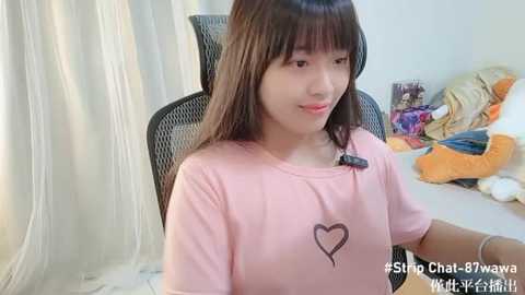 Media: Video of an Asian woman with straight black hair, wearing a pink shirt with a heart symbol, sitting in a black office chair, surrounded by colorful stuffed animals in a bright room.