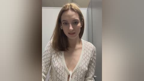 Media: A video of a young Caucasian woman with light skin and straight, shoulder-length blonde hair, wearing a white, patterned cardigan with a deep V-neck, standing in a dimly lit, beige-walled hallway.