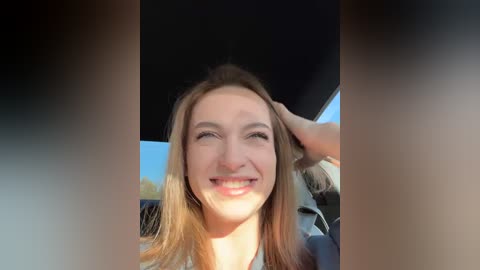 Media: Video of a smiling young woman with light skin and straight, shoulder-length blonde hair, sitting in a car with a black interior. She is wearing a light-colored top. The background outside shows a clear blue sky.