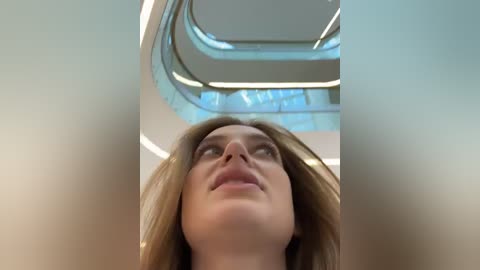 Media: A video of a Caucasian woman with long, light brown hair, looking upward with a neutral expression, captured from a low angle in a modern, indoor space with curved, light blue-tinted ceiling panels and bright, ambient lighting.