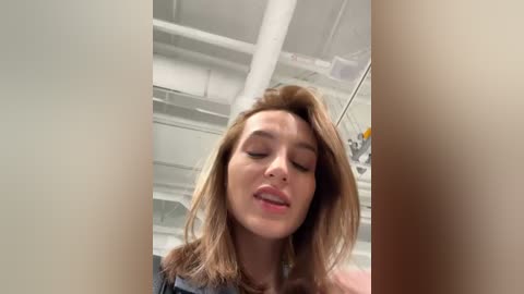 Media: A video of a young woman with light brown hair, wearing a black top, smiling slightly, captured from a low angle, showing off the ceiling's white ductwork and industrial lighting.