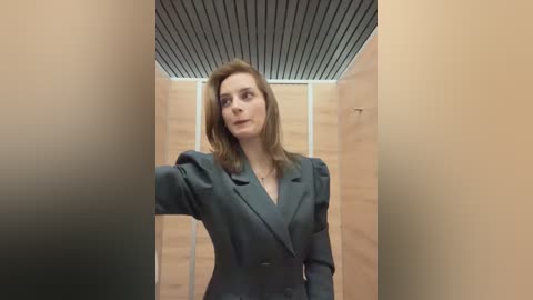 Media: Video of a Caucasian woman with shoulder-length brown hair, wearing a dark green blazer, standing in a narrow, beige-walled hallway with a slanted, dark ceiling. She appears thoughtful.