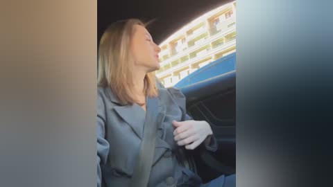 Media: Video of a blonde woman in a gray coat, driving a car with a blue seatbelt on, looking to the right, with a multi-story building in the background.