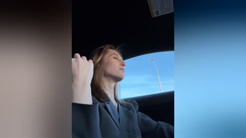 Media: Video of a woman with fair skin and shoulder-length blonde hair, wearing a blue blazer, gazing out a car window at a clear blue sky.