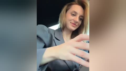 Media: Video of a smiling, fair-skinned woman with straight, shoulder-length blonde hair in a grey blazer, holding a phone, seated in a car.