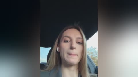 Media: A video of a young woman with fair skin, straight light brown hair, and light makeup, smiling slightly, wearing a black jacket, inside a car.