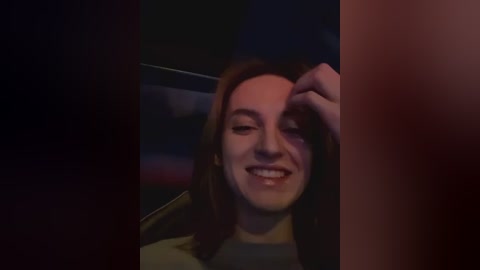 Media: A video of a young woman with fair skin, long brown hair, and a broad smile, captured in a dimly lit car interior.