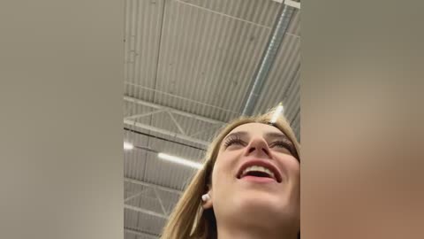 Media: A video of a smiling woman with blonde hair, wearing glasses, standing in an industrial building with a corrugated metal ceiling and exposed pipes.