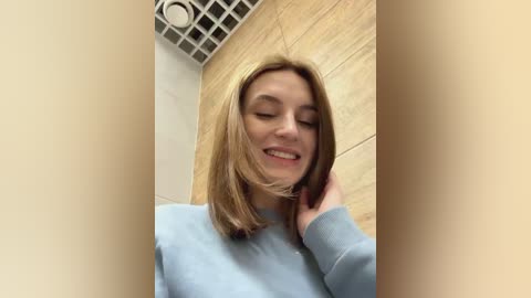 Media: Video of a young Caucasian woman with light brown hair and a light blue sweater, smiling, with her hair blowing in an indoor setting featuring beige tiles and a grid ceiling.