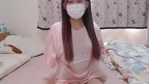 Media: Video of an East Asian woman with long, straight, dark brown hair, wearing a pink \"I Love NY\" t-shirt and a white face mask, sitting on a bed in a cluttered, dimly lit room.