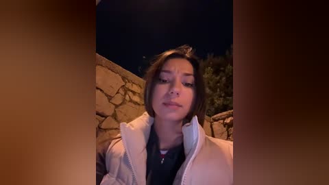 Media: Video of a young Asian woman with straight black hair, fair skin, and a slight smile, wearing a beige jacket, standing outdoors at night beside a stone wall, with a dark background.