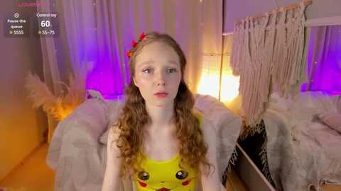 Media: Video of a young girl with fair skin and long, curly brown hair adorned with a red flower, wearing a Pikachu tank top, sitting on a plush, gray couch in a cozy, dimly lit room with purple and orange ambient lighting.