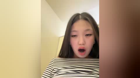 Media: Video of a young East Asian woman with long black hair, wearing a striped shirt, looking surprised and open-mouthed, standing in a narrow, beige-walled hallway with a wooden door in the background.