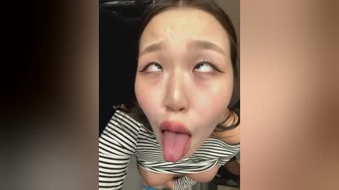 Media: Video of an Asian woman with light skin, dark hair, and wearing a striped top, sticking out her tongue provocatively. The background is blurred, focusing on her face.