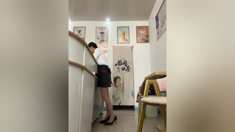 Media: A video of a woman in a white blouse and black skirt, leaning against a white wall, in a minimalist, brightly lit room with framed posters and a Japanese sign.