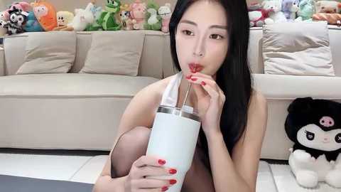 Media: Video of an East Asian woman with long black hair, wearing red nail polish, drinking from a large clear cup, sitting on a white floor in a plush room with stuffed toys and a beige sofa.