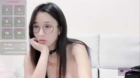 Media: A video of an East Asian woman with long black hair and glasses, wearing a black choker, leaning on a white couch in a minimalistic room with a calendar and clock visible.