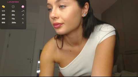 Media: Video of a young woman with fair skin, long black hair, and a white off-shoulder top, looking contemplative in a dimly lit room with a closed door and a blurred background.