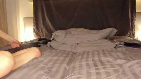 Media: Video of a person lying on a bed with a striped gray blanket, holding a black remote, and a dark curtain behind them.