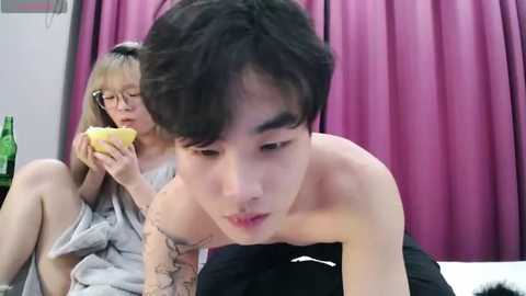 Media: Video of an Asian man with short black hair, shirtless, eating a banana, tattooed arm. Behind him, an Asian woman in glasses, wearing a gray dress, sits on a bed with pink curtains.