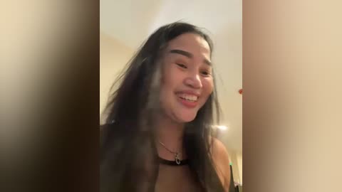 Media: A video of an Asian woman with long black hair, smiling widely, wearing a black choker and a light-colored top, in a blurred, beige-colored room.