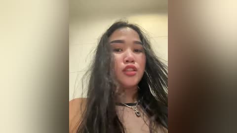 Media: A video of a young Asian woman with long, dark hair, wearing a black choker and silver earrings, standing in an indoor setting with beige walls.