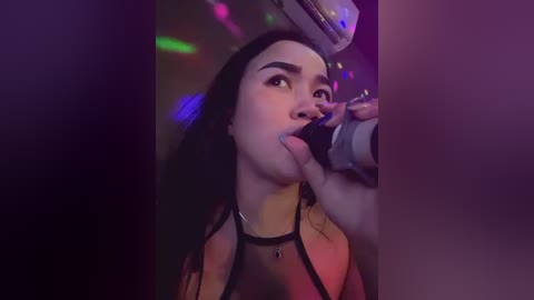 Media: Video of a young woman with long black hair, wearing a black halter top, licking an ice cream cone while inside a dimly lit, colorful, disco-themed venue.