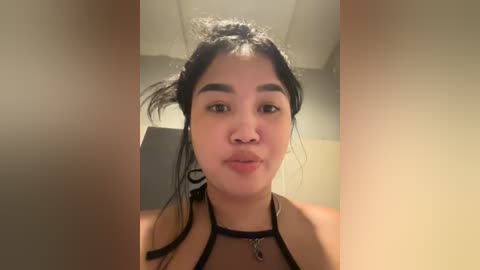 Media: A video of a young Asian woman with long, black hair tied in a messy bun. She wears a black halter top and stands in a dimly lit room with beige walls.