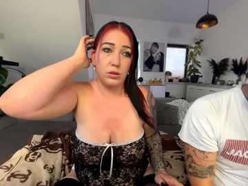 Media: Video of a plus-size woman with long red hair, large breasts, and tattoos, wearing a black lace corset, sitting on a bed in a modern, minimalistic bedroom with black and white decor.