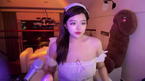 Media: Video of a young Asian woman with long dark hair in a maid outfit, holding a drink, surrounded by plush toys in a dimly lit room.