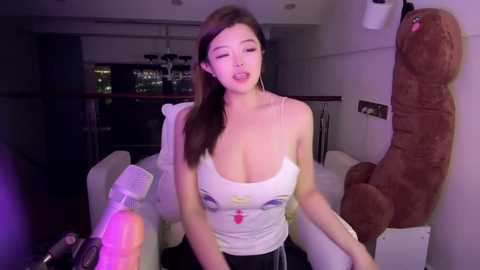 Media: Video of an Asian woman with long brown hair, wearing a white tank top, leaning against a brown plush toy in a dimly lit, modern living room.