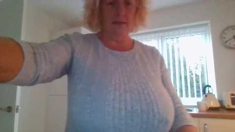 Media: Video of a middle-aged woman with curly blonde hair, wearing a light blue knitted sweater, standing in a bright, modern kitchen with white walls, a window, and a clock on the wall.