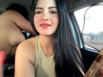 Media: Video of a Latina woman with long black hair, wearing a beige tank top, smiling, in a car, with blurred figures in the background.