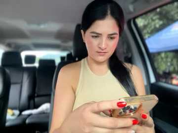 Media: Video of a young woman with long black hair, fair skin, wearing a beige sleeveless top, sitting in a car, texting on her phone with red-painted nails.