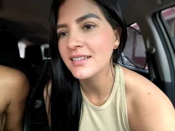 Media: Video of a smiling woman with long black hair, wearing a sleeveless beige top, sitting in a car. Background shows blurred car interior.